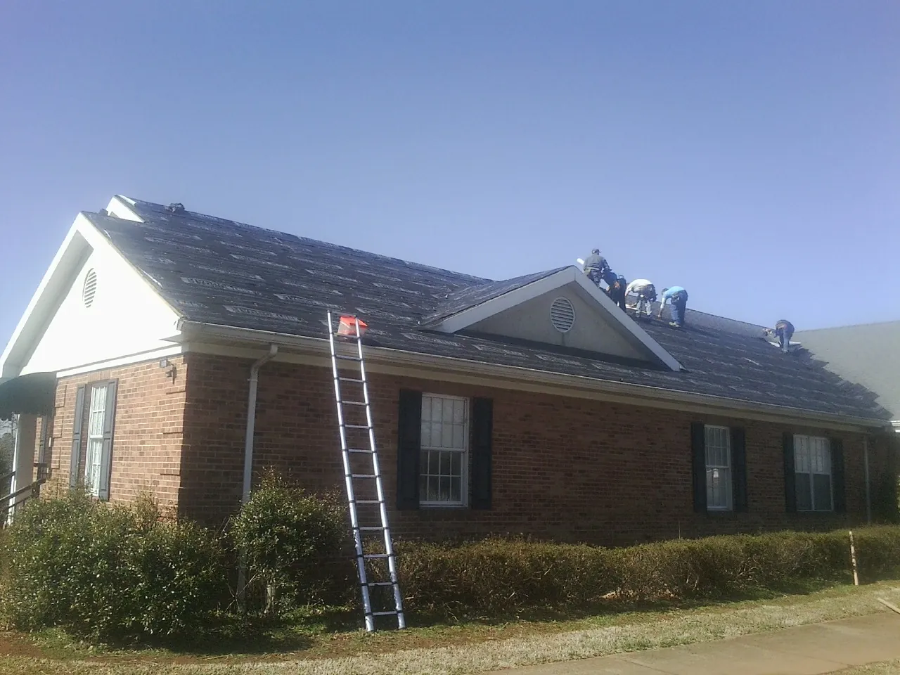 Crew tearing shingles off a brick ranch roof with a ladder set — Metal Roof Repair work by our crew in Magna