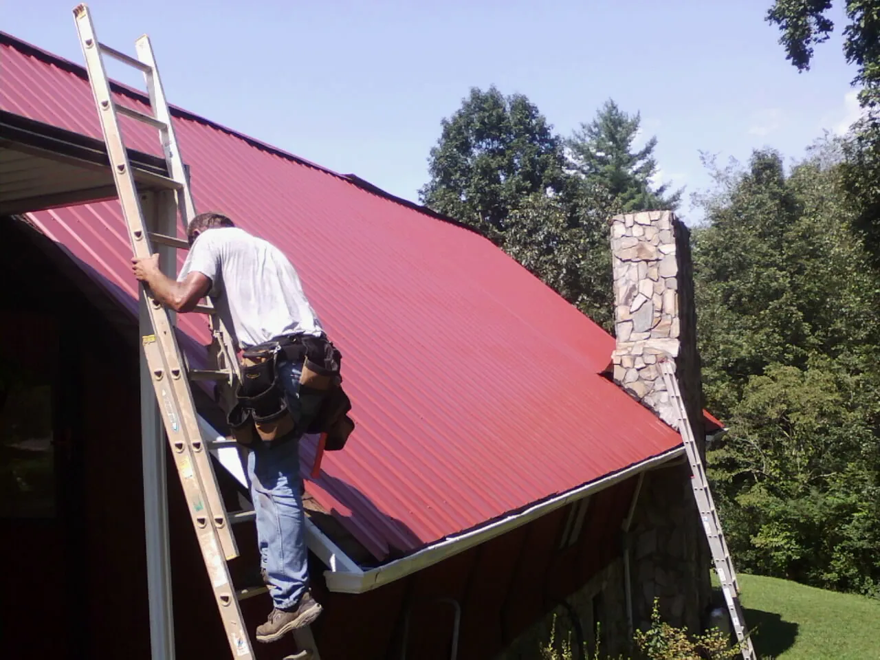 Roofer on a ladder beside a red metal roof and stone chimney — Soffit & Fascia Repair by Roofing Brothers in Magna
